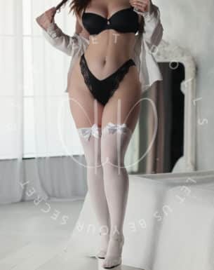 Woman modeling in black lingerie (bra and lace panties) with white thigh-high stockings and heels in a bright bedroom setting.