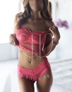 Woman modeling pink lace lingerie, lifting her top to reveal matching panties in a softly lit indoor setting (face blurred).