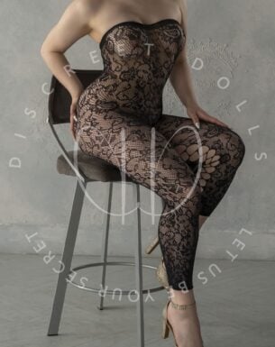 Woman seated on a bar stool wearing a black lace strapless bodysuit and gold heels, posing for a lingerie shoot.