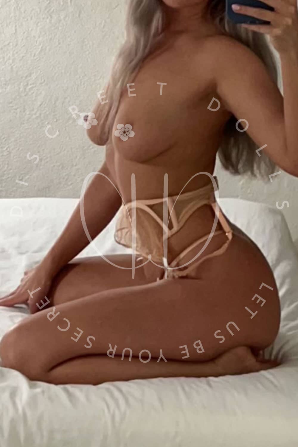 Topless woman kneeling on a bed, wearing beige strappy lingerie, taking a mirror selfie.